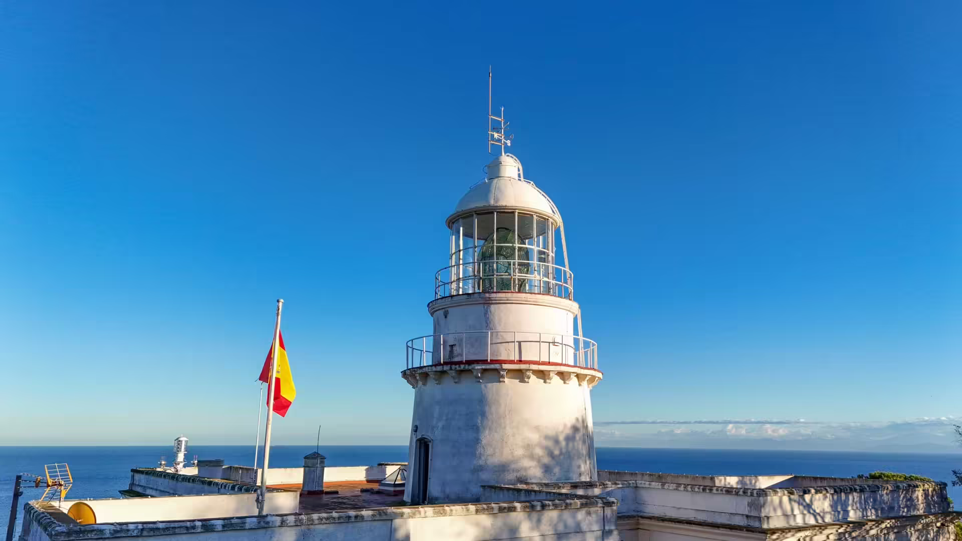 Biela veža majáka Faro de Punta Almina so španielskou vlajkou a Gibraltárskym prielivom v pozadí