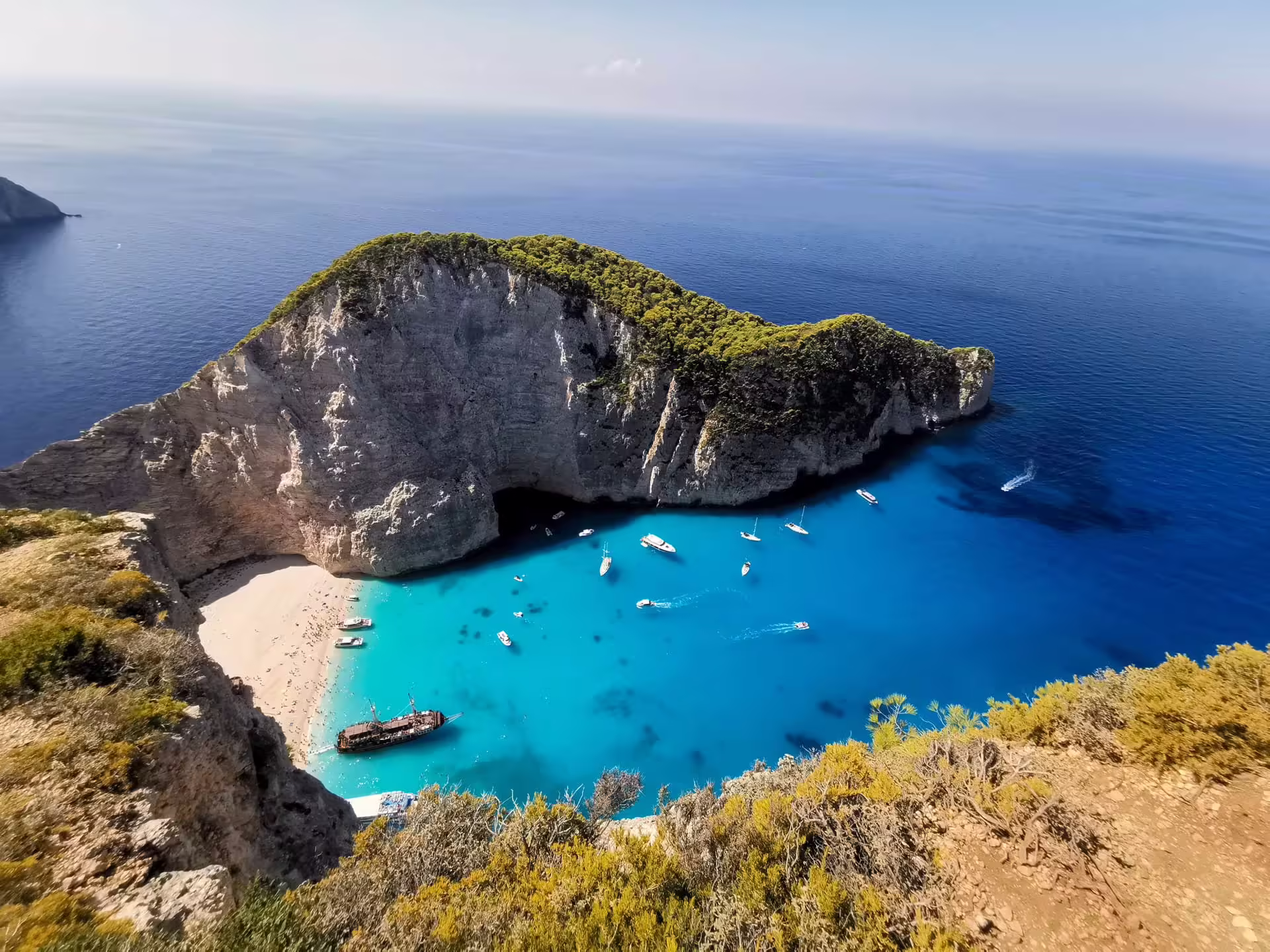 Pláž Navagio z výšky, vrak lode Panagiotis na svetlom brehu, vysoké skalné steny a turistické lode v tyrkysovej vode