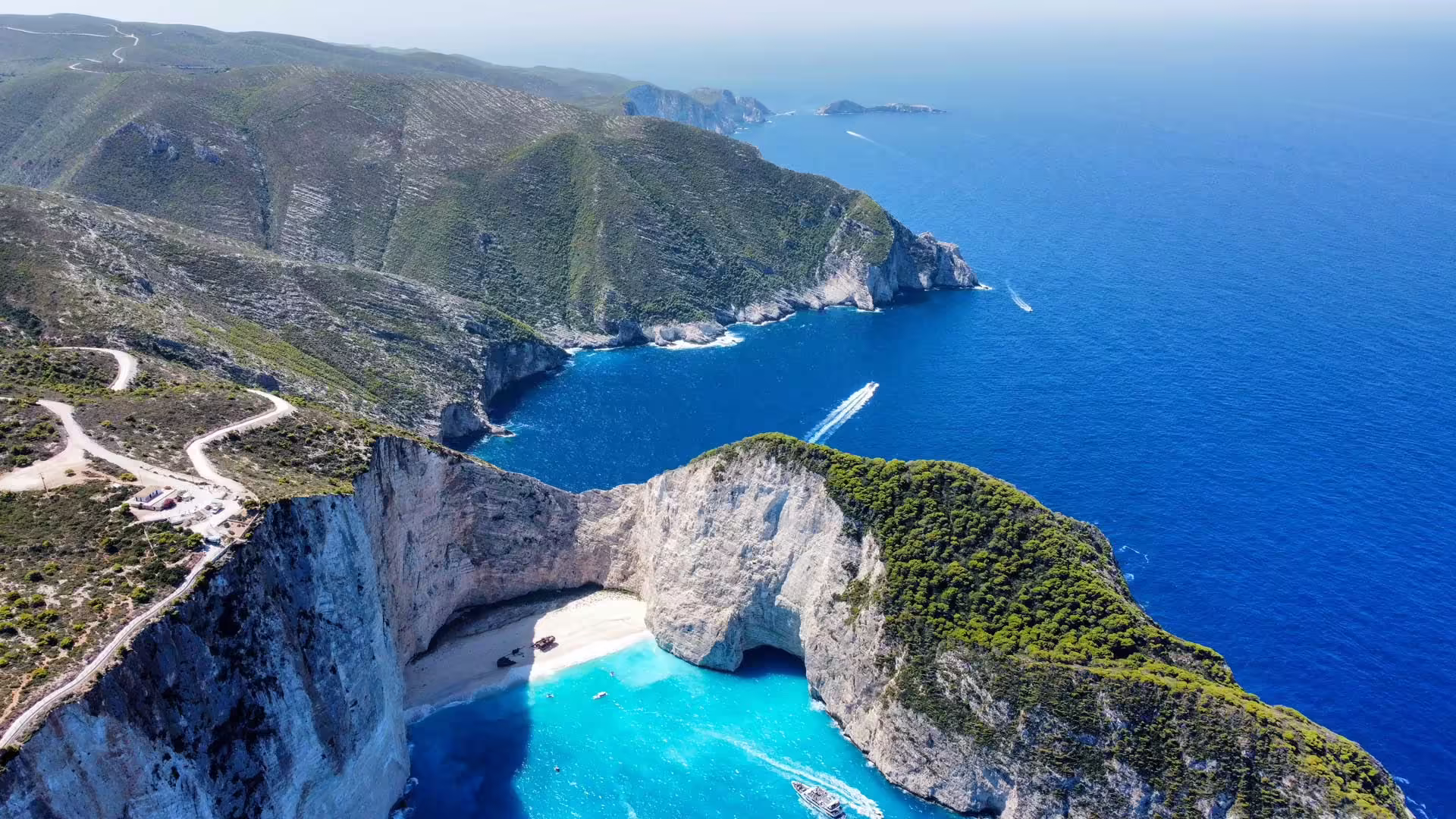 Letecký pohľad na zátoku Navagio s modrým Iónskym morom, vápencovými útesmi, malou plážou a kľukatou cestou nad pobrežím