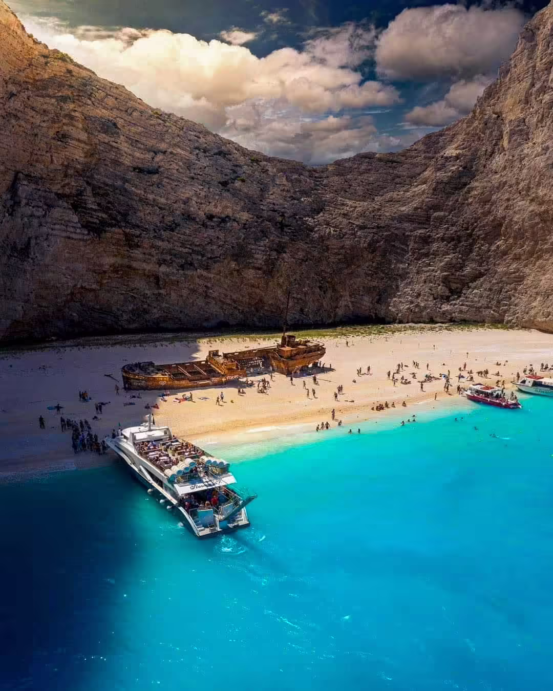 Turisti na svetlej pláži Navagio medzi vysokými vápencovými útesmi, hrdzavý vrak Panagiotis a výletná loď pri brehu